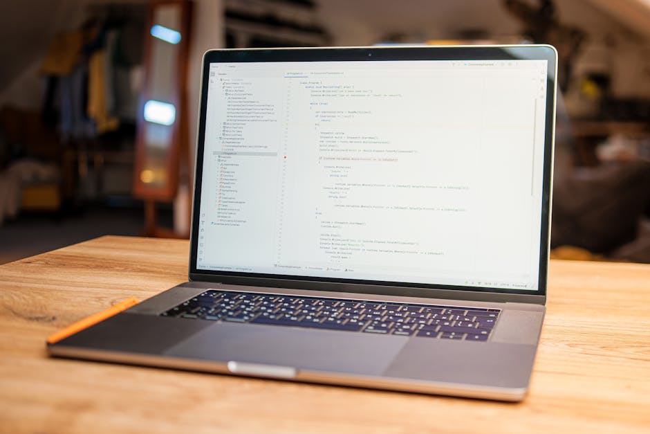 A close-up view of a person's hands working on a laptop computer placed on a dark surface indoors. The screen displays lines of code with colorful syntax highlighting against a predominantly black bac